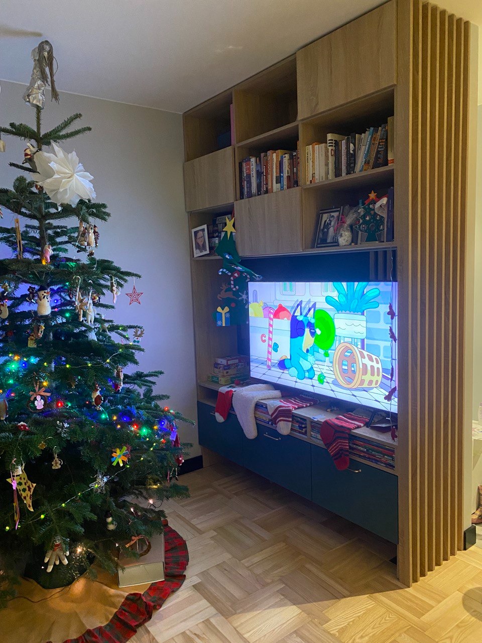 Wieloelementowa zabudowa meblowa z drewnianymi lamelami po prawej stronie, z telewizorem w środkowej części i półkami z książkami u góry, w pokoju stoi choinka z ozdobami świątecznymi.