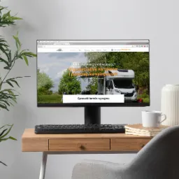 Monitor wyświetla stronę internetową wypożyczalni kamperów z widokiem kampera na tle zieleni, na biurku obok stoi biała filiżanka i wazon