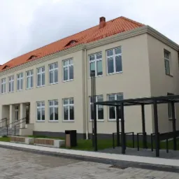 Elewacja dwukondygnacyjnego budynku z czerwonym dachem, białymi oknami i nowoczesną dobudówką ze szkła i metalu, widoczne elementy zagospodarowania terenu: ławki, kosze, stojaki na rowery.