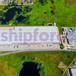 Widok z lotu ptaka na rozległy kompleks magazynowy z rampami załadunkowymi, otoczony zielenią i zbiornikami wodnymi, z widocznym logo firmy na dachu.