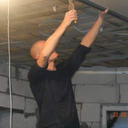 Bud Isa - Mężczyzna montuje metalowy profil sufitowy za pomocą szczypiec, widoczne kable i konstrukcja sufitu podwieszanego, ściany z białych bloczków.