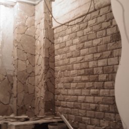 Bud Isa - Fragment ściany w trakcie wykańczania: część pokryta kamienną okładziną, część z imitacją cegły oraz przygotowana powierzchnia do malowania. Widoczne narzędzia i pozostałości po pracach.