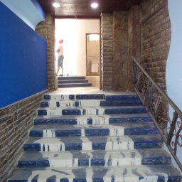 Bud Isa - Schody z niebieskimi stopnicami pokryte świeżo nałożoną, mokrą gładzią, widoczny fragment ściany z cegły oraz dekoracyjny sufit z drewnianą boazerią i elementami niebieskiej sztukaterii, w tle...