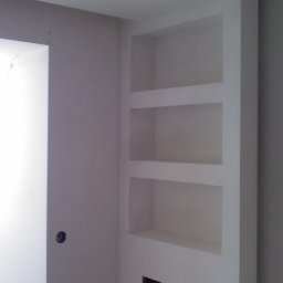 Bud Isa - Wnęka z półkami w ścianie po remoncie, widoczny fragment grzejnika w dolnej części wnęki, ściany pomalowane na biało, surowy stan wykończenia.