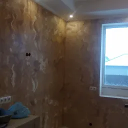 Ściana w pokoju pokryta dekoracyjną, brązowo-białą farbą imitującą marmur, widoczne gniazdka elektryczne, skrzynka elektryczna, okno i fragment wyposażenia.