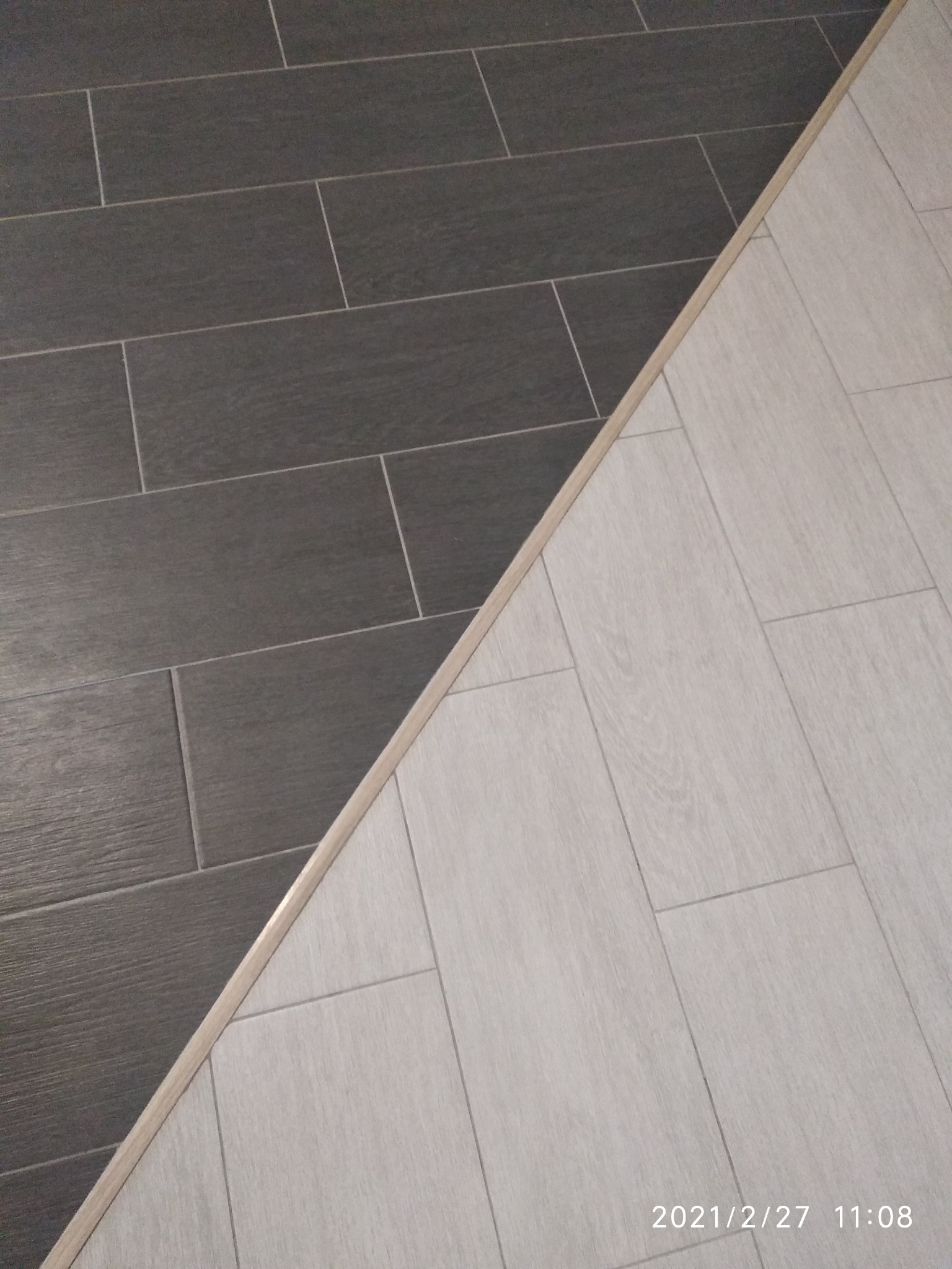 Połączenie dwóch rodzajów płytek podłogowych: ciemnoszarych, drewnopodobnych płytek oraz jasnych, drewnopodobnych płytek, oddzielonych listwą progową w kolorze drewna.