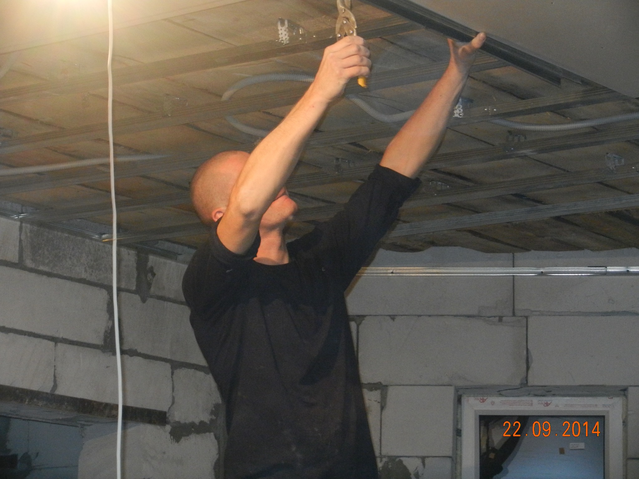 Mężczyzna montuje metalowy profil sufitowy za pomocą szczypiec, widoczne kable i konstrukcja sufitu podwieszanego, ściany z białych bloczków.