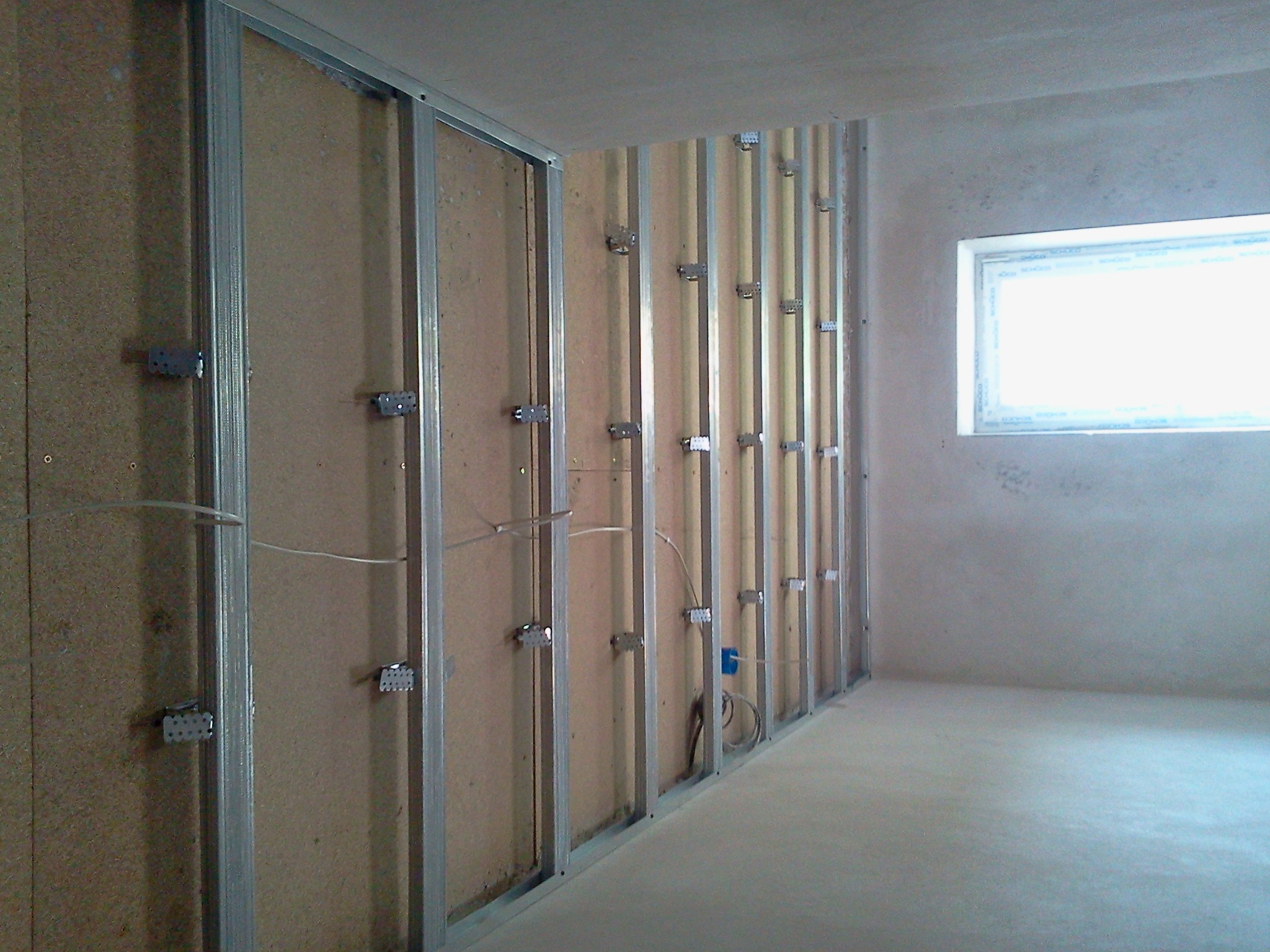 Konstrukcja szkieletowa z profili stalowych i płyt OSB, widoczne przewody elektryczne, przygotowanie do montażu okładzin ściennych w pomieszczeniu z oknem.