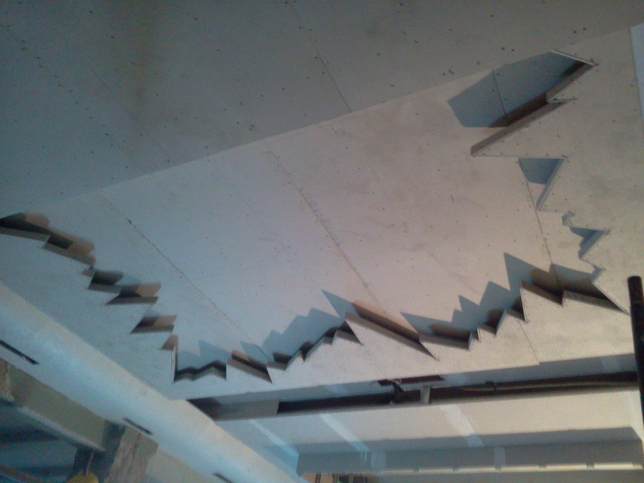 Niedokończony sufit z płyt kartonowo-gipsowych z widocznymi metalowymi profilami, nieregularne otwory w kształcie zębów piły, widoczne elementy konstrukcyjne budynku.