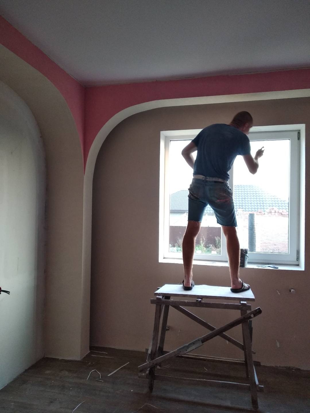 Mężczyzna w krótkich spodenkach stoi na drewnianym podeście przy oknie, poprawiając wykończenie ściany w pomieszczeniu z łukowatym przejściem i pomalowanym na różowo sufitem.