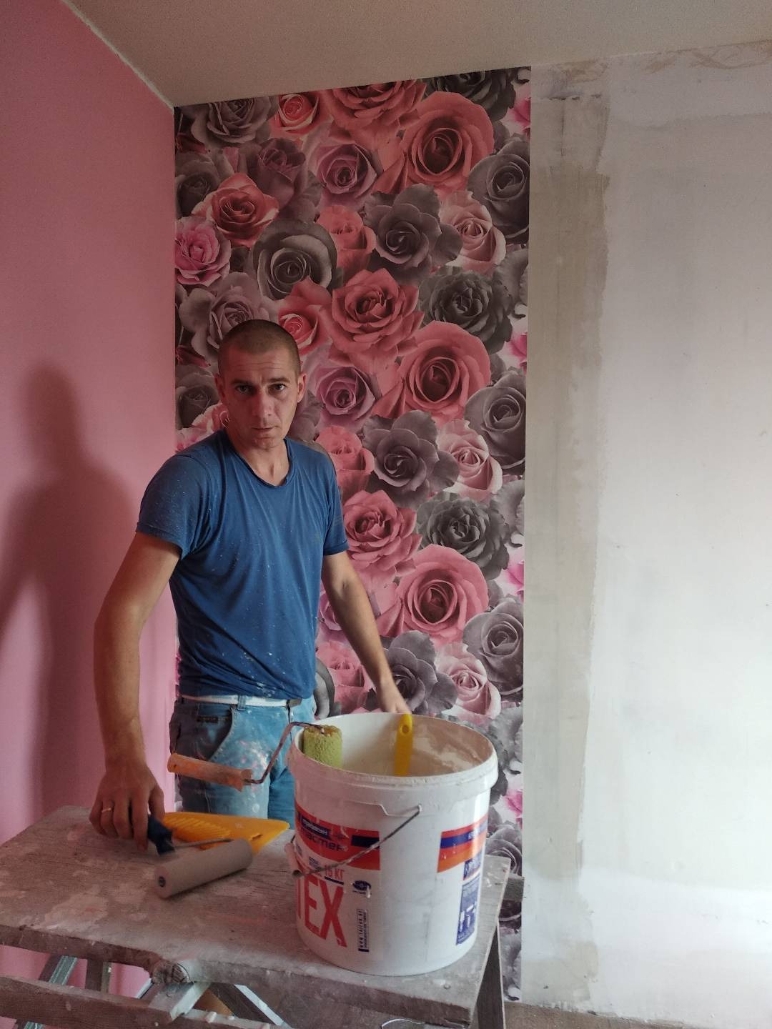 Pracownik w Kłodzku podczas tapetowania ściany w kwiaty, widoczny wałek malarski i wiadro z klejem na stole malarskim, obok surowa ściana przygotowana do dalszych prac.