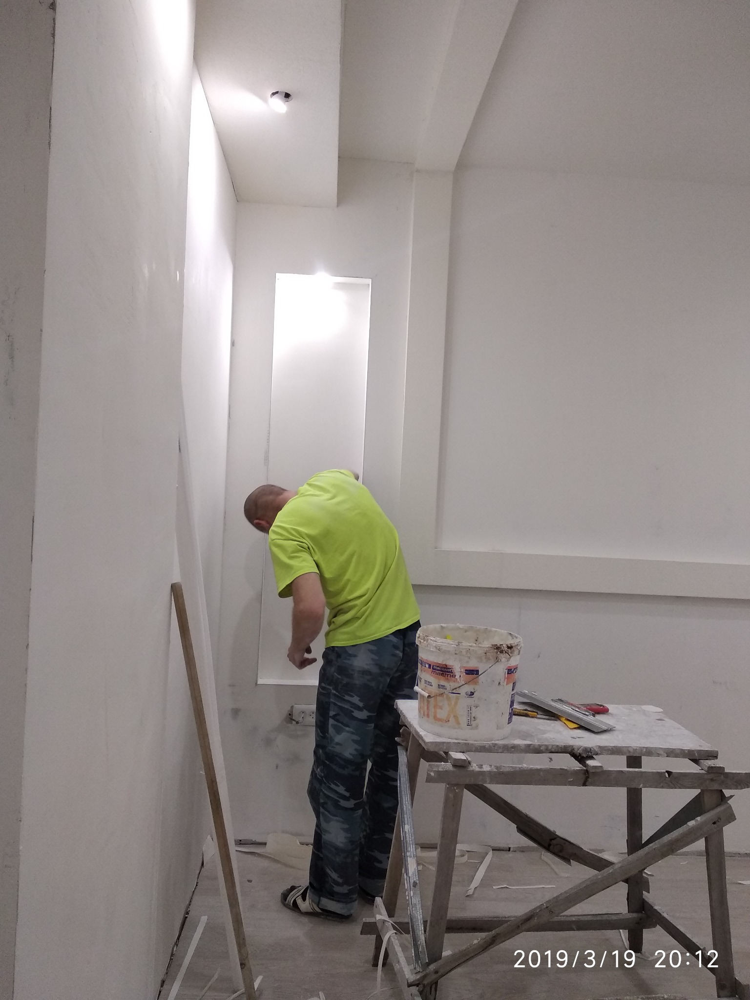 Pracownik w neonowej koszulce szpachluje ścianę w pomieszczeniu z wnęką, obok stoi drabina malarska z wiadrem farby i narzędziami.