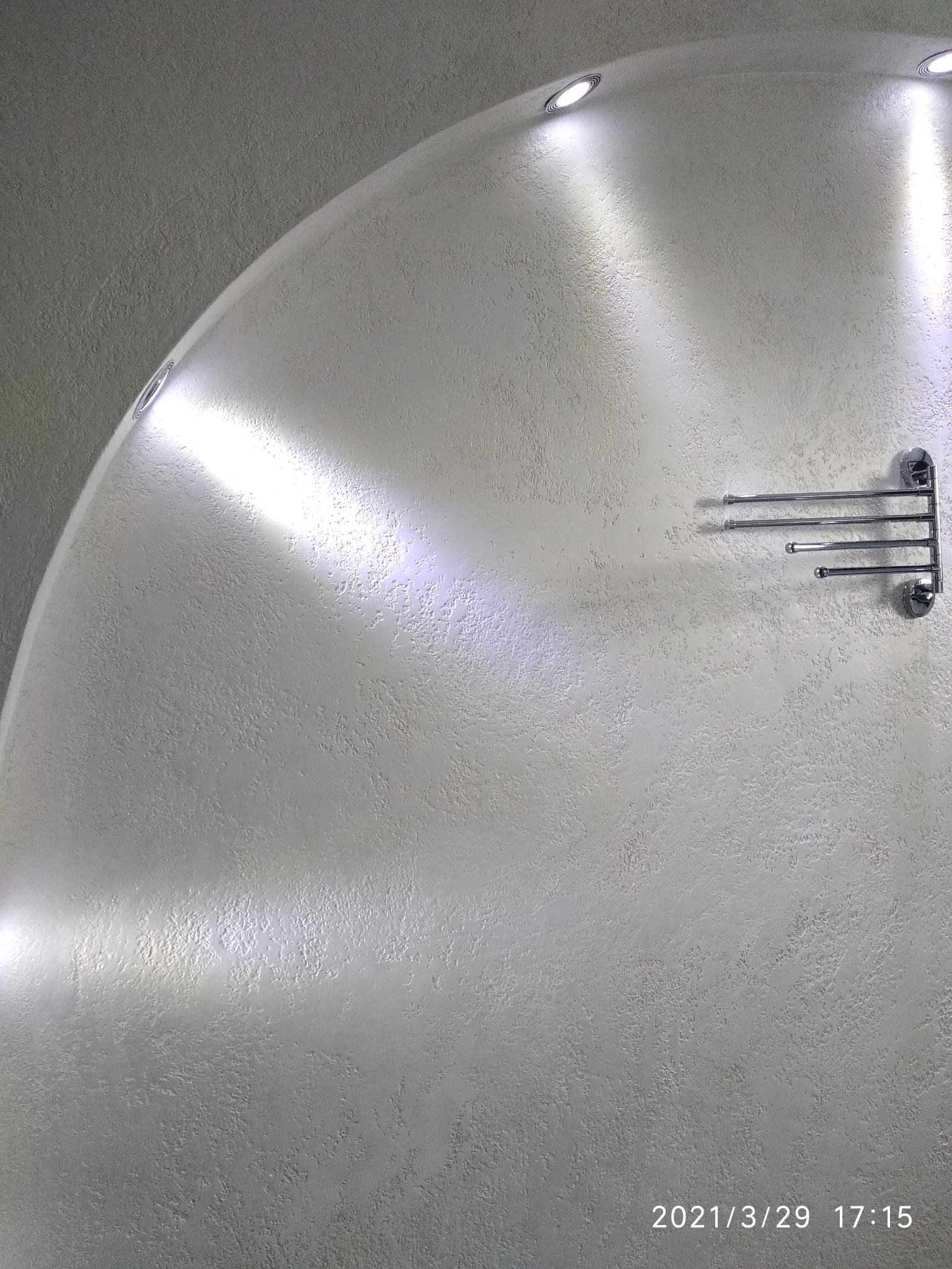 Ściana łazienki wykończona dekoracyjną strukturą, oświetlona punktowymi lampami sufitowymi i wyposażona w chromowany wieszak na ręczniki.