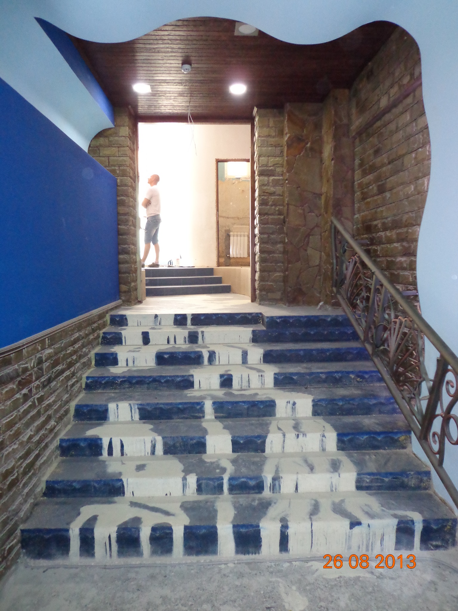 Schody z niebieskimi stopnicami pokryte świeżo nałożoną, mokrą gładzią, widoczny fragment ściany z cegły oraz dekoracyjny sufit z drewnianą boazerią i elementami niebieskiej sztukaterii, w tle...