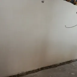 Świeżo nałożona, gładka, jasnoszara ściana wewnętrzna z widocznymi śladami po szpachlowaniu, niedokończona instalacja elektryczna w górnym rogu oraz surowy beton na podłodze.