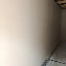 Świeżo otynkowana ściana w surowym wnętrzu, widoczne elementy konstrukcji sufitu i przewody przy podłodze.