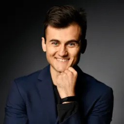 Uśmiechnięty mężczyzna w granatowej marynarce, z dłonią pod brodą, na ciemnoszarym tle.