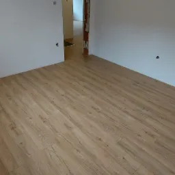 Jasny, drewnopodobny laminat ułożony w nowym pokoju z białymi ścianami. Widoczne niedokończone gniazdka elektryczne i taśma miernicza na parapecie.