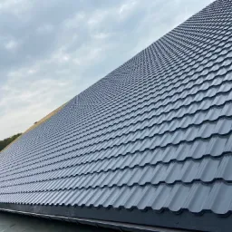 Nowy, grafitowy dach z blachy trapezowej z widocznym fragmentem rynny i lasu w tle, sfotografowany pod kątem.