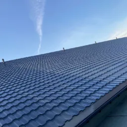 Nowy, ciemny dach z metalowej dachówki modułowej, widok z dołu, z kominkami wentylacyjnymi, na tle błękitnego nieba z smugą kondensacyjną