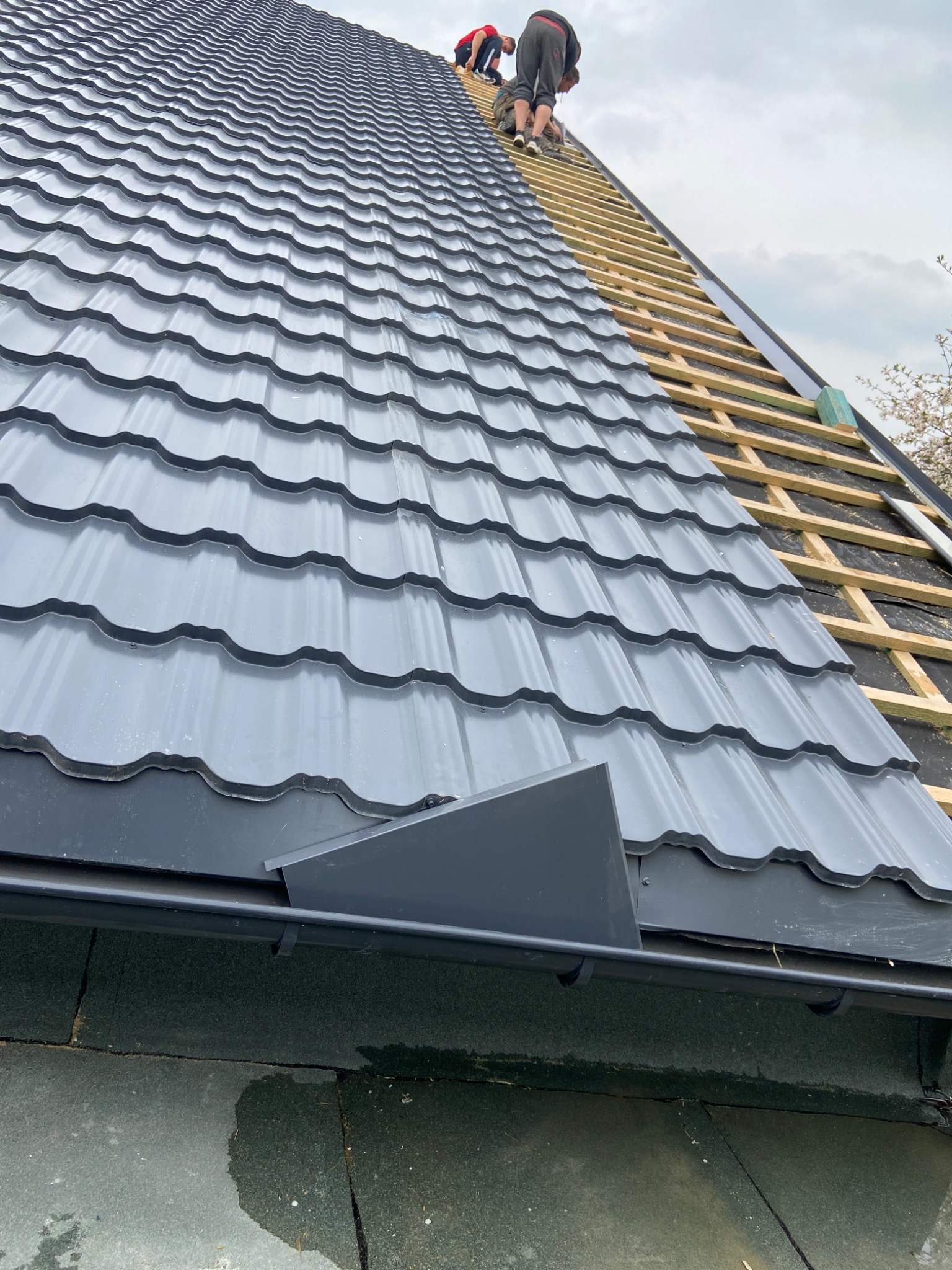 Montaż szarego pokrycia dachowego z widocznymi elementami drewnianej konstrukcji i pracownikami na dachu, perspektywa z dołu.