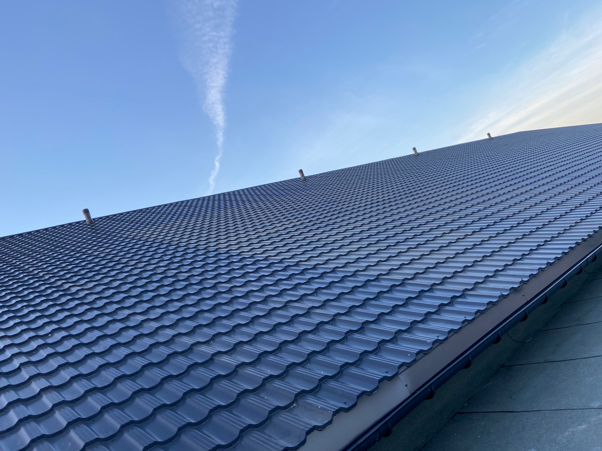 Nowy, ciemny dach z metalowej dachówki modułowej, widok z dołu, z kominkami wentylacyjnymi, na tle błękitnego nieba z smugą kondensacyjną
