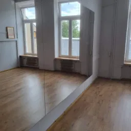 Pusta sala taneczna z dużym lustrem na ścianie, parkietem, grzejnikami pod oknami i lampą kulą na suficie.