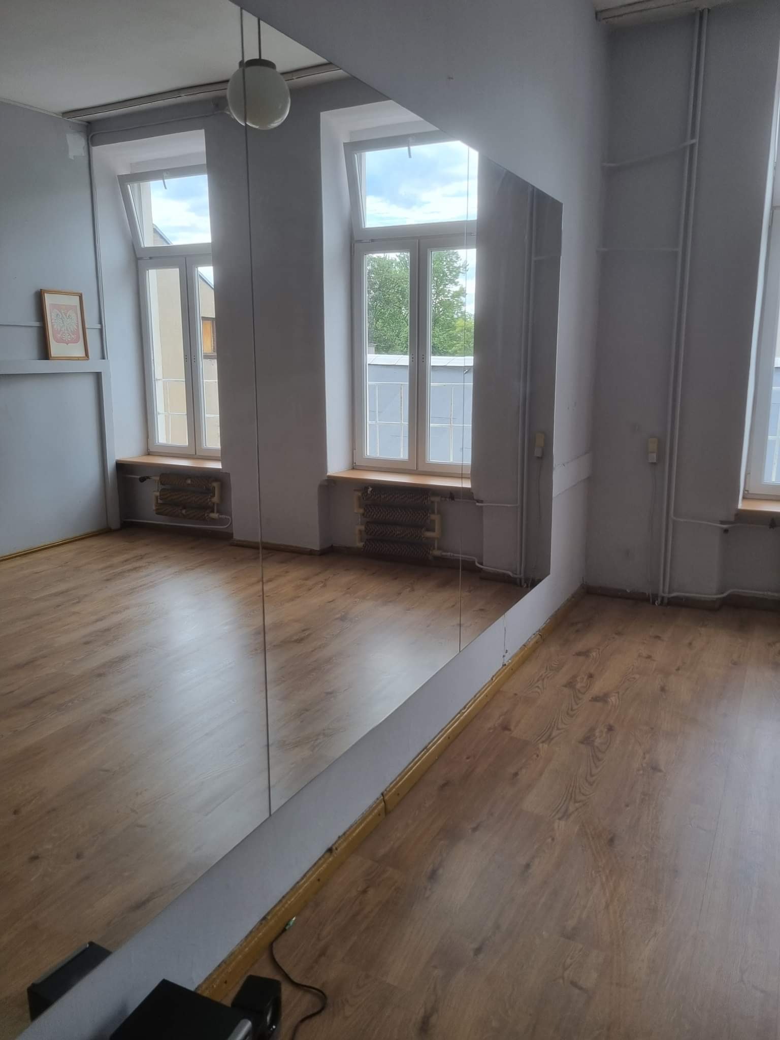 Pusta sala taneczna z dużym lustrem na ścianie, parkietem, grzejnikami pod oknami i lampą kulą na suficie.