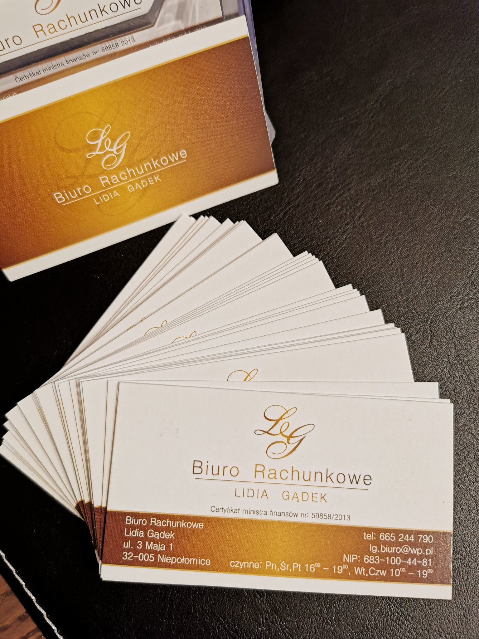 Stos wizytówek biura rachunkowego Lidia Gądek, z adresem, numerem telefonu i NIP, leżący na ciemnym blacie.