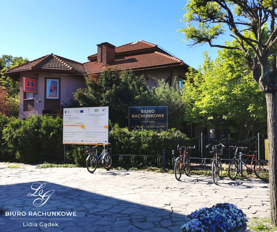 Budynek biura rachunkowego z szyldem informacyjnym, rowerami przed wejściem i tablicą informacyjną o dofinansowaniu unijnym, widok w słoneczny dzień.