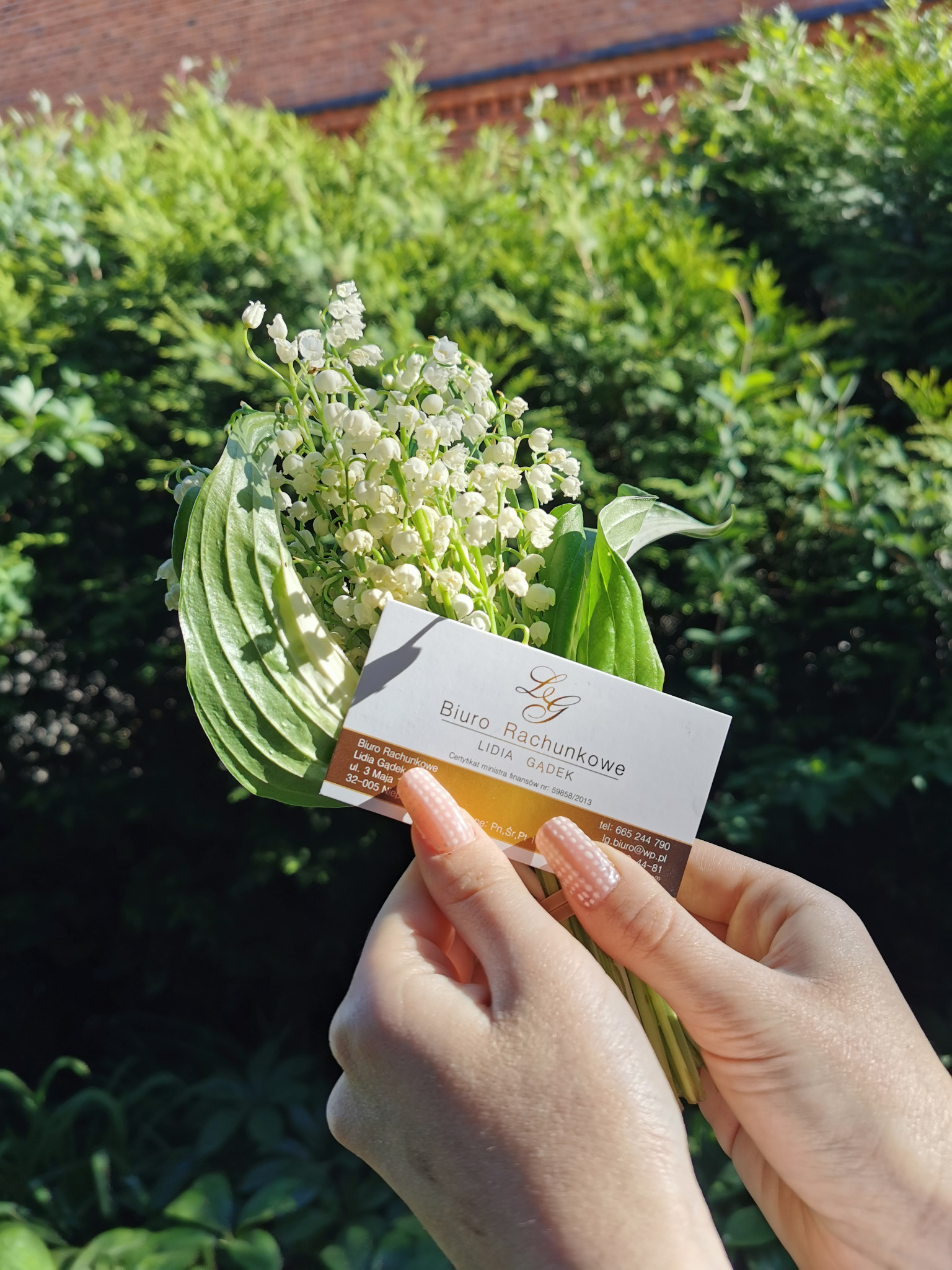 Dłonie trzymają bukiecik konwalii z wizytówką biura rachunkowego Lidia Gądek na tle zieleni.