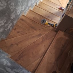 Zakład budowlano-remontowy TOM - Wykończone drewniane schody z narzędziami (szlifierka kątowa) i akcesoriami (silikon) na stopniach. Widoczna ściana z tapetą w kwiaty i fragment buta.