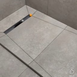 Zakład budowlano-remontowy TOM - Beżowe płytki podłogowe w łazience z odpływem liniowym. Dwie kulki, pomarańczowa i biała, leżą na płytkach. Widok z góry, fragment podłogi i ściany.