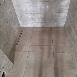 Zakład budowlano-remontowy TOM - Wykończona kabina prysznicowa z odpływem liniowym, wyłożona płytkami w odcieniach szarości i beżu. Widok z góry, ukazujący precyzję ułożenia płytek i estetykę wykończenia.