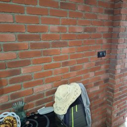 Zakład budowlano-remontowy TOM - Ściana z czerwonej cegły z widocznymi fugami, fragment czarnego włącznika światła, stół z przedmiotami w dolnej części kadru. Dekoracyjna ściana z cegły.