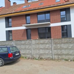 Zakład budowlano-remontowy TOM - Nowoczesny dom jednorodzinny z czerwoną cegłą na elewacji, czarnymi oknami i balkonem, widziany zza betonowego ogrodzenia. Na pierwszym planie zaparkowany samochód.