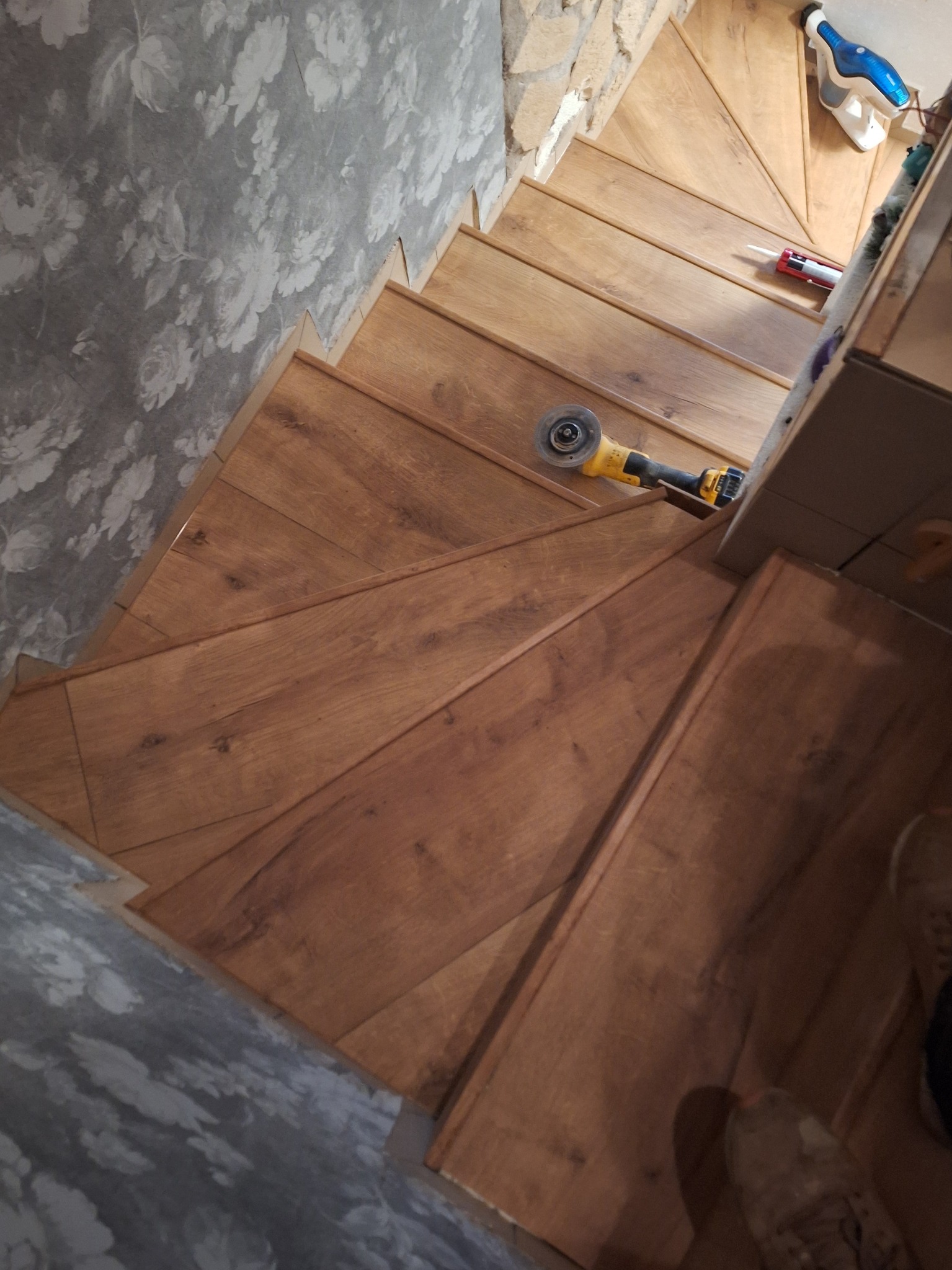 Wykończone drewniane schody z narzędziami (szlifierka kątowa) i akcesoriami (silikon) na stopniach. Widoczna ściana z tapetą w kwiaty i fragment buta.