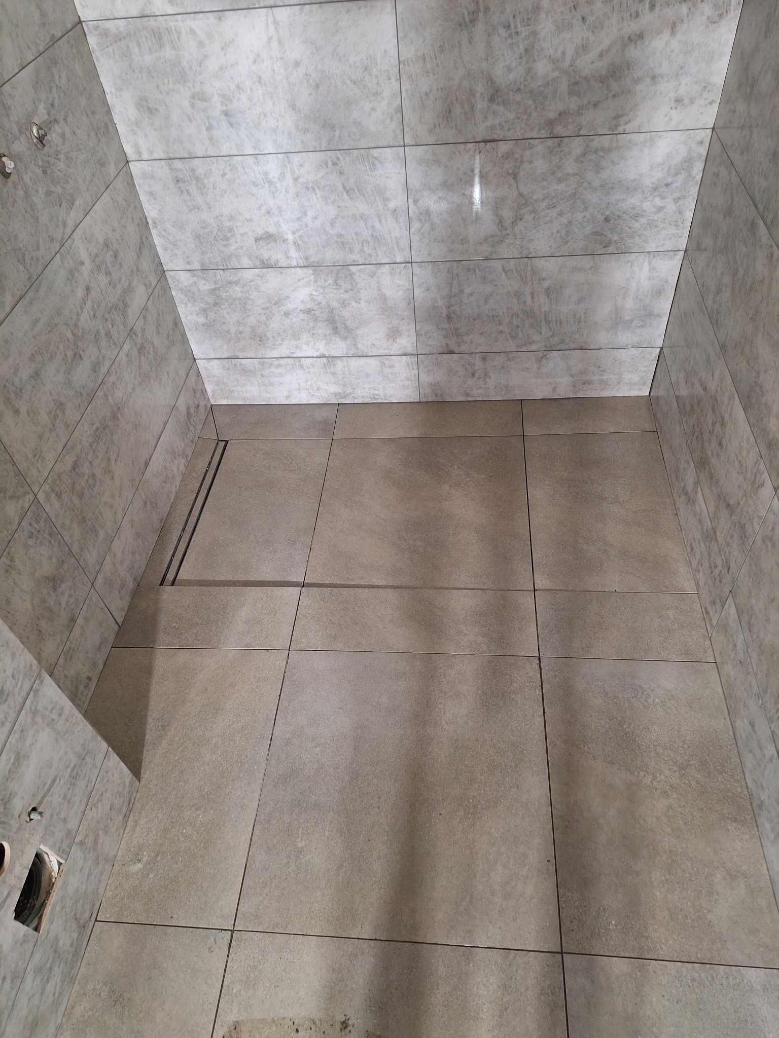 Wykończona kabina prysznicowa z odpływem liniowym, wyłożona płytkami w odcieniach szarości i beżu. Widok z góry, ukazujący precyzję ułożenia płytek i estetykę wykończenia.