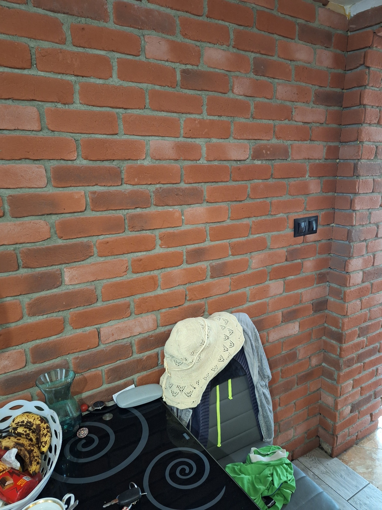 Ściana z czerwonej cegły z widocznymi fugami, fragment czarnego włącznika światła, stół z przedmiotami w dolnej części kadru. Dekoracyjna ściana z cegły.