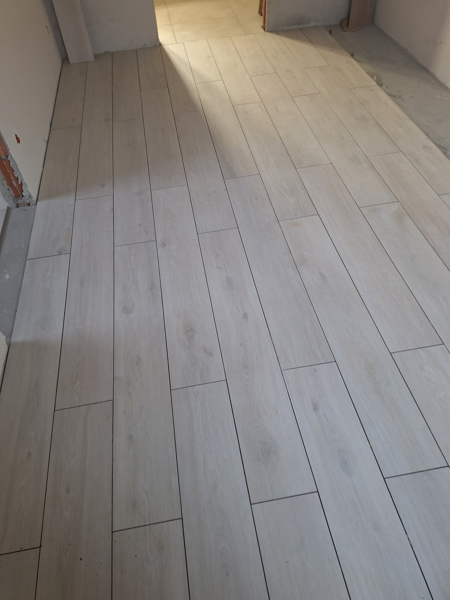 Ułożone panele podłogowe w jasnym odcieniu drewna, widok z góry, z delikatnym cieniem padającym z góry. Widoczne fugi między panelami i fragment ściany w tle.