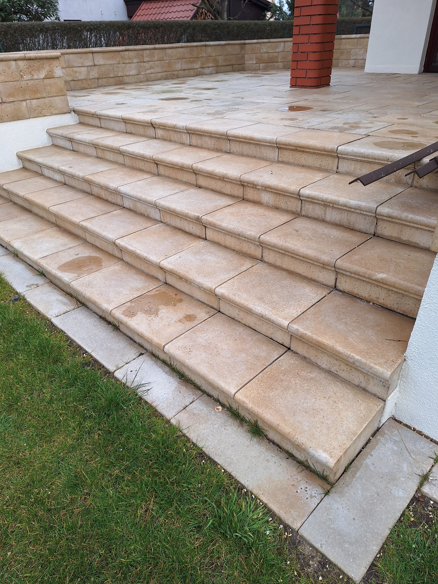 Schody zewnętrzne wyłożone jasnymi, kwadratowymi płytkami z delikatnym zaokrągleniem krawędzi, prowadzące do budynku z ceglanym filarem i zielonym trawnikiem.