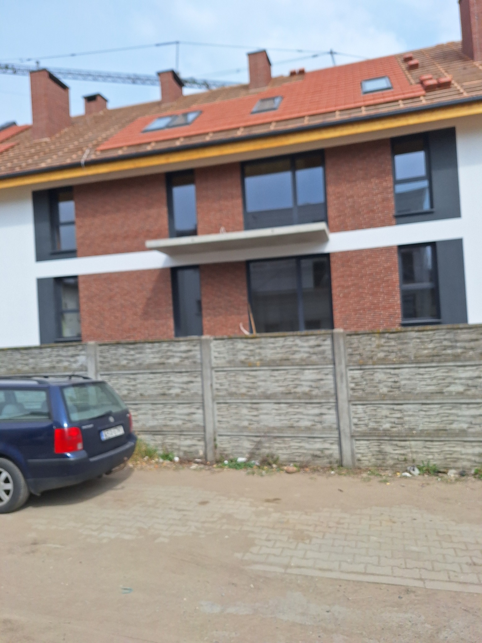 Nowoczesny dom jednorodzinny z czerwoną cegłą na elewacji, czarnymi oknami i balkonem, widziany zza betonowego ogrodzenia. Na pierwszym planie zaparkowany samochód.
