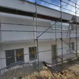 Świeżo otynkowana elewacja domu jednorodzinnego z widocznymi rusztowaniami, zabezpieczonymi oknami i drzwiami folią malarską oraz fragmentami gruzu u podstawy.