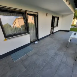 Biała elewacja domu z czarnymi ramami okiennymi i drzwiami, szara, kamienna posadzka tarasu, widoczny fragment ogrodu z rusztowaniami.