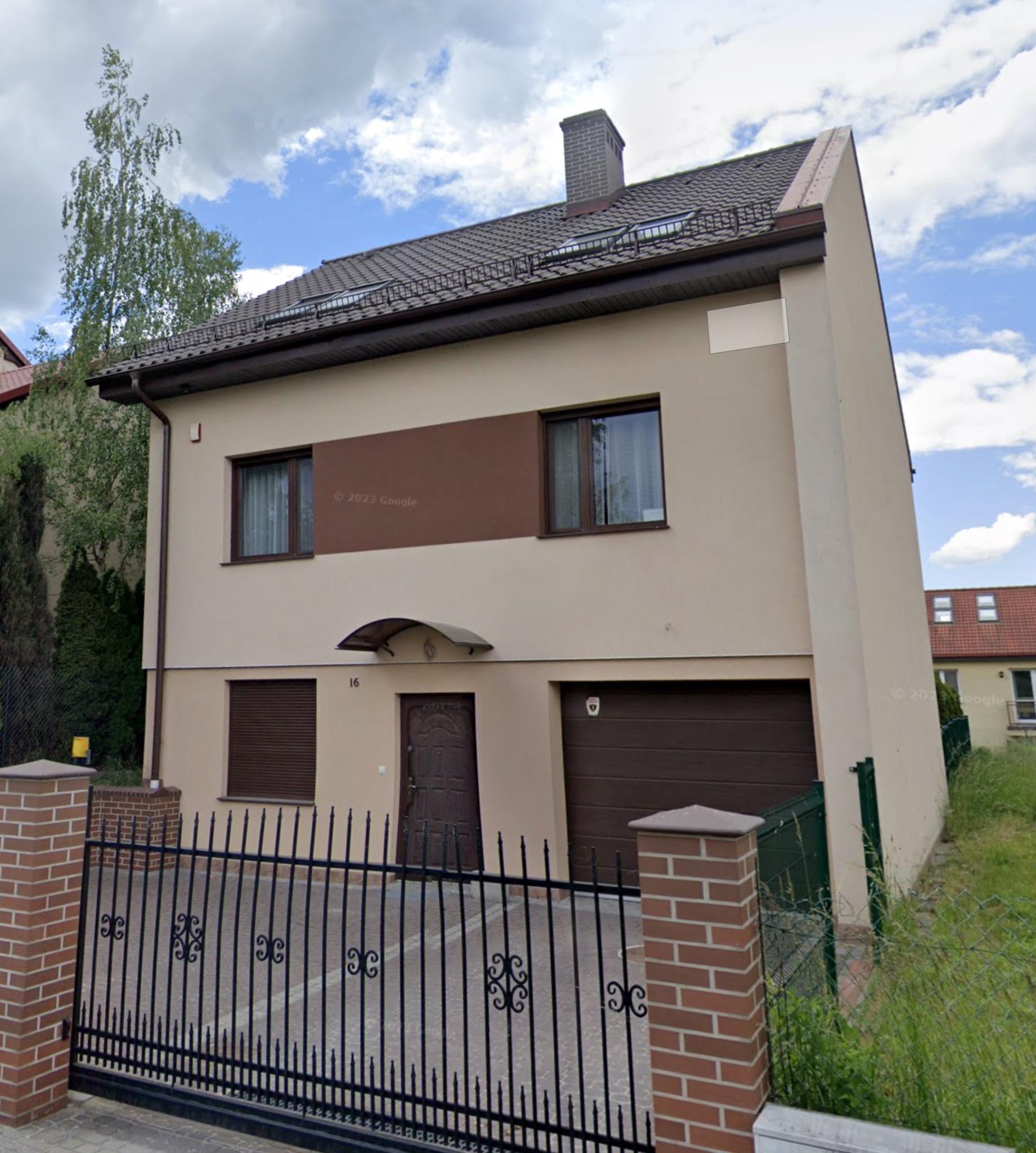 Dwukondygnacyjny dom jednorodzinny z brązowymi akcentami, widoczny z zewnątrz przez czarne, ozdobne ogrodzenie.