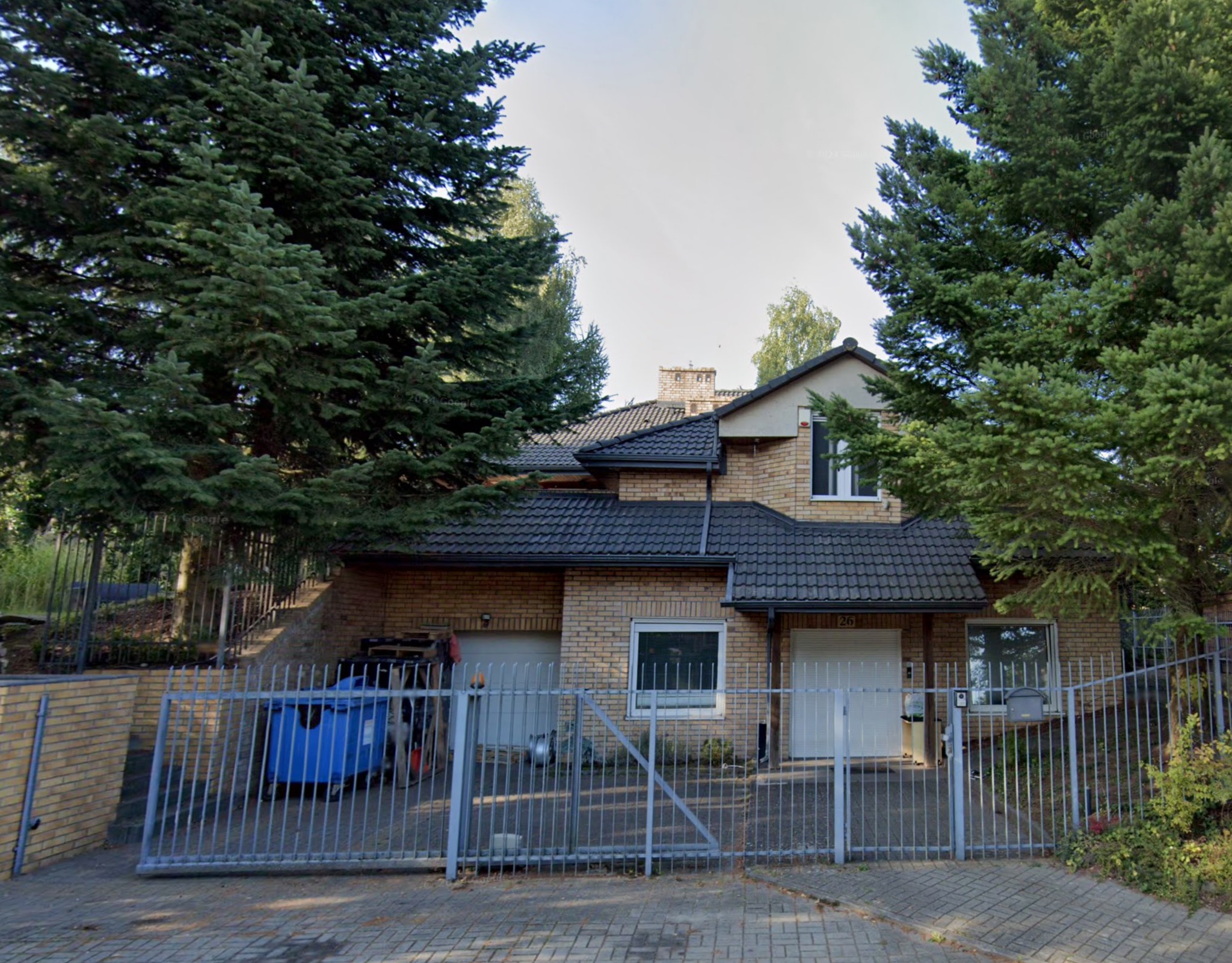 Dwukondygnacyjny dom jednorodzinny z garażami, elewacją z jasnej cegły, ciemnym dachem i stalowym ogrodzeniem, częściowo zasłonięty przez wysokie drzewa iglaste.