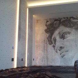 Juliusz Łękawski - Ściana z dekoracyjną fakturą, oświetleniem LED w pionowych szczelinach i dużym muralem z wizerunkiem Dawida na sąsiedniej ścianie, zabezpieczona folią malarską na podłodze, widoczne stopy w obuwiu...