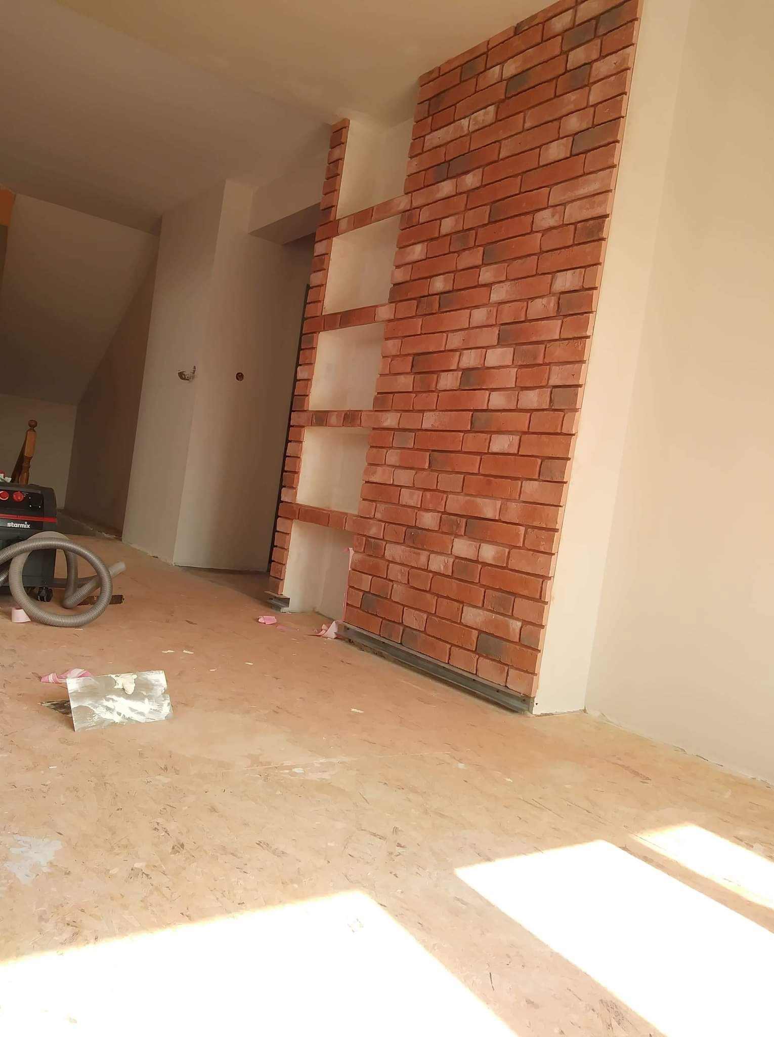 Fragment ściany z cegły klinkierowej w trakcie montażu z wnękami, widoczny odkurzacz przemysłowy i narzędzia na podłodze przygotowanej do dalszych prac wykończeniowych.