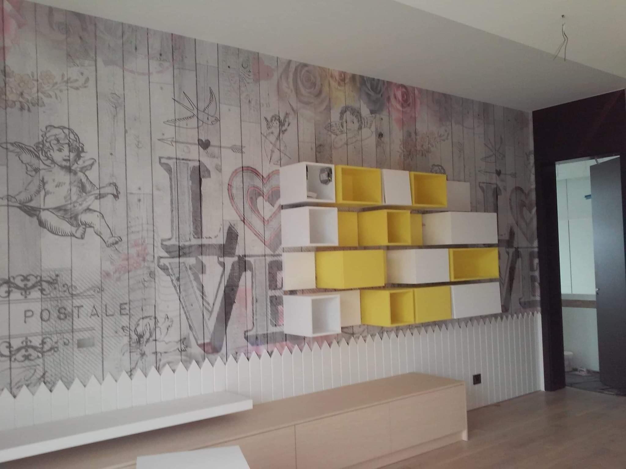 Pokój z fototapetą w stylu vintage z motywem anioła i napisu LOVE, uzupełniony białymi i żółtymi modułowymi półkami oraz niską szafką RTV.