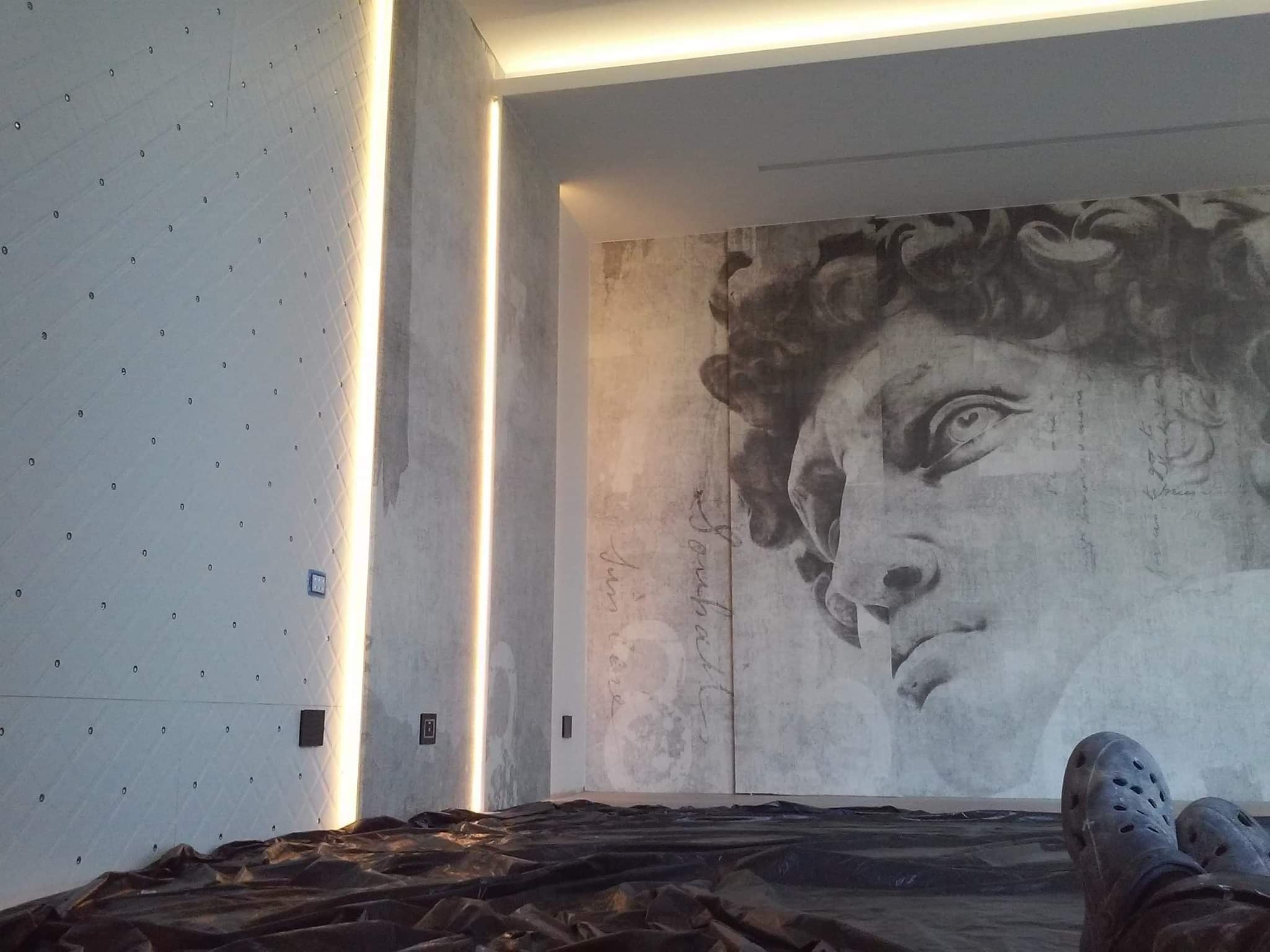 Ściana z dekoracyjną fakturą, oświetleniem LED w pionowych szczelinach i dużym muralem z wizerunkiem Dawida na sąsiedniej ścianie, zabezpieczona folią malarską na podłodze, widoczne stopy w obuwiu...